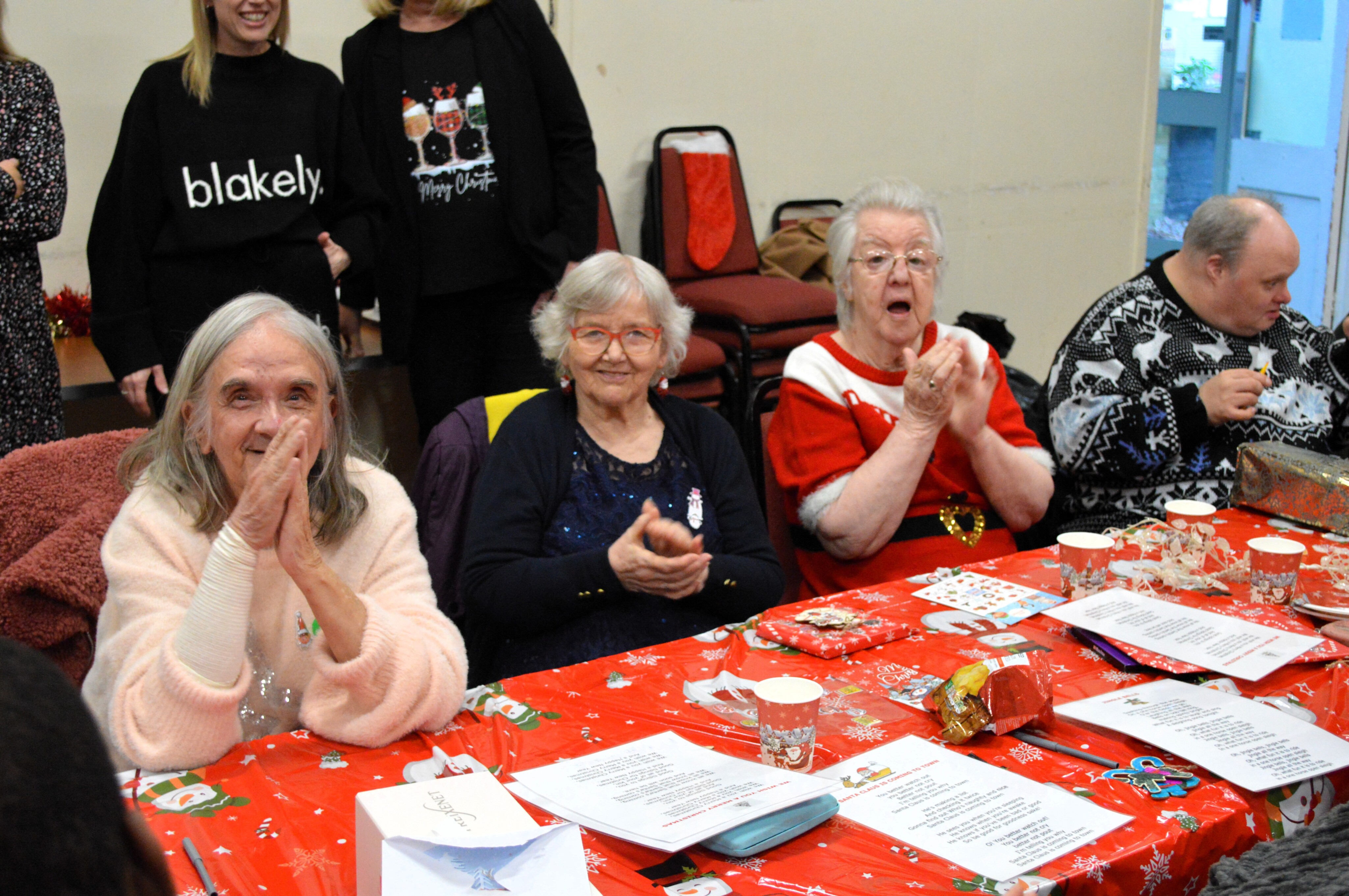 Group of people sitting around a table decorated with Christmas-themed items and papers, engaging in a festive activity indoors.