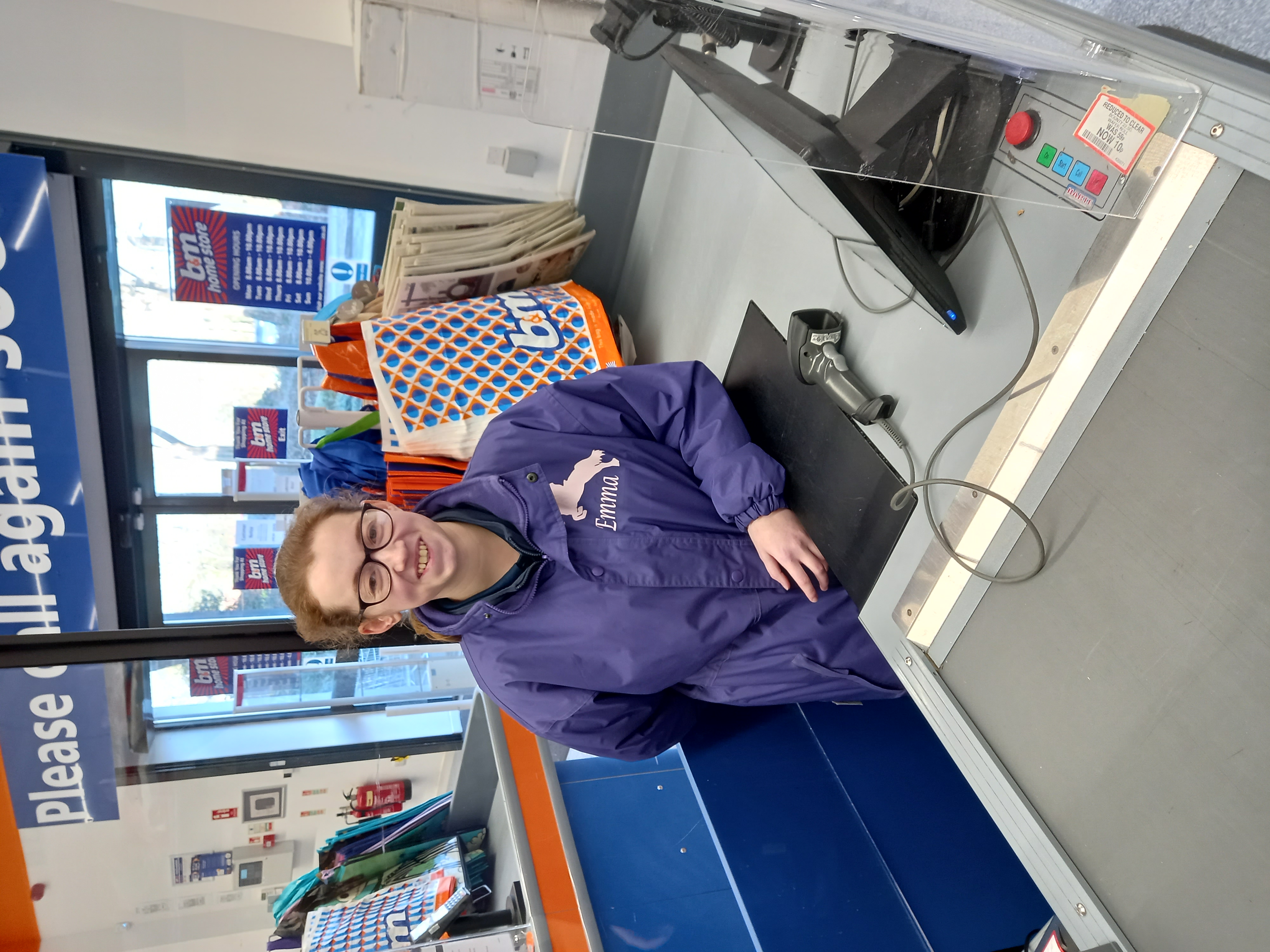 Emma wearing a purple jacket stands behind a checkout counter with a barcode scanner and monitor in a store.