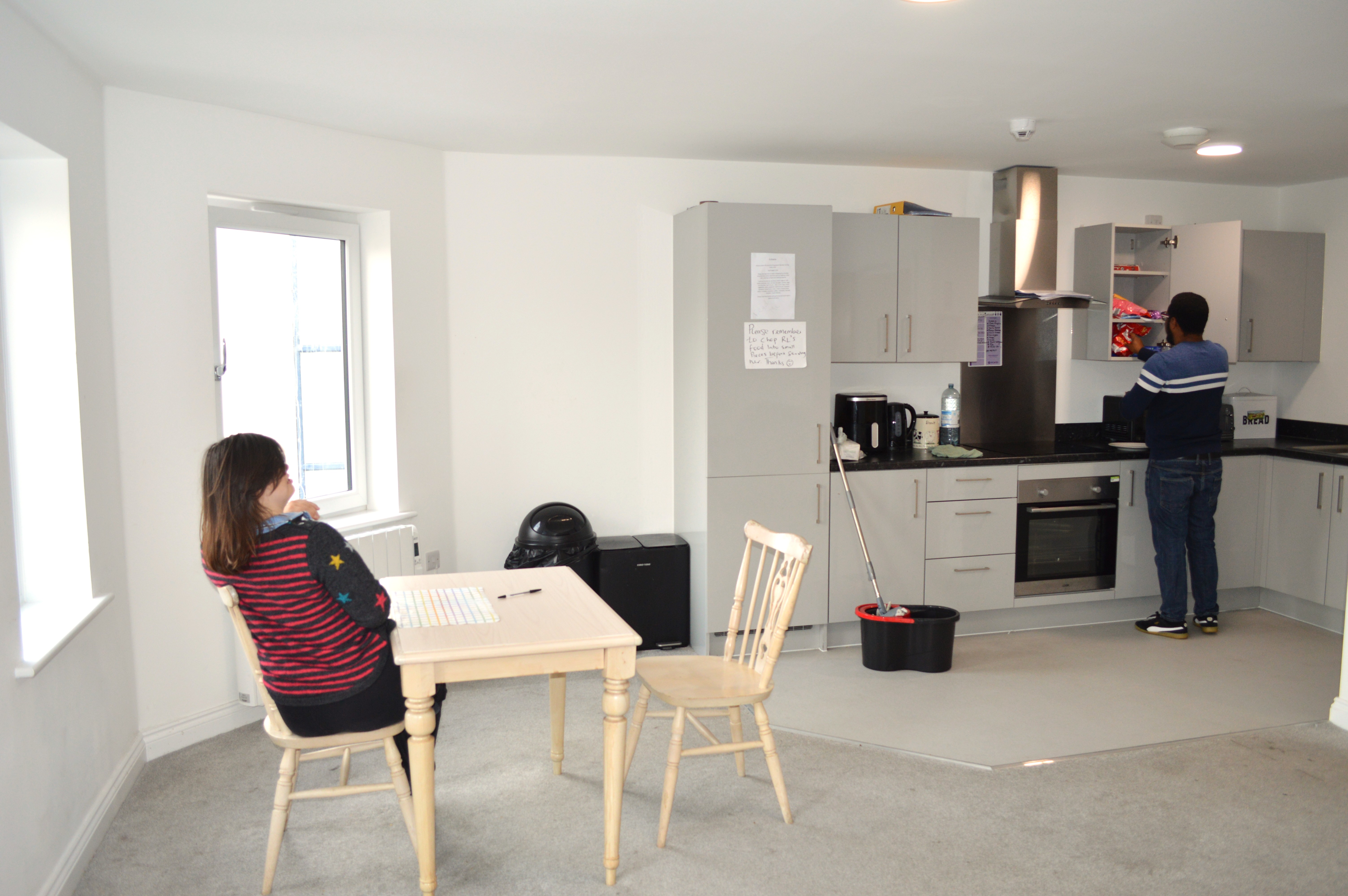 Rhi sits at the table whilst Anthony looks in the cupboard in the kitchen.