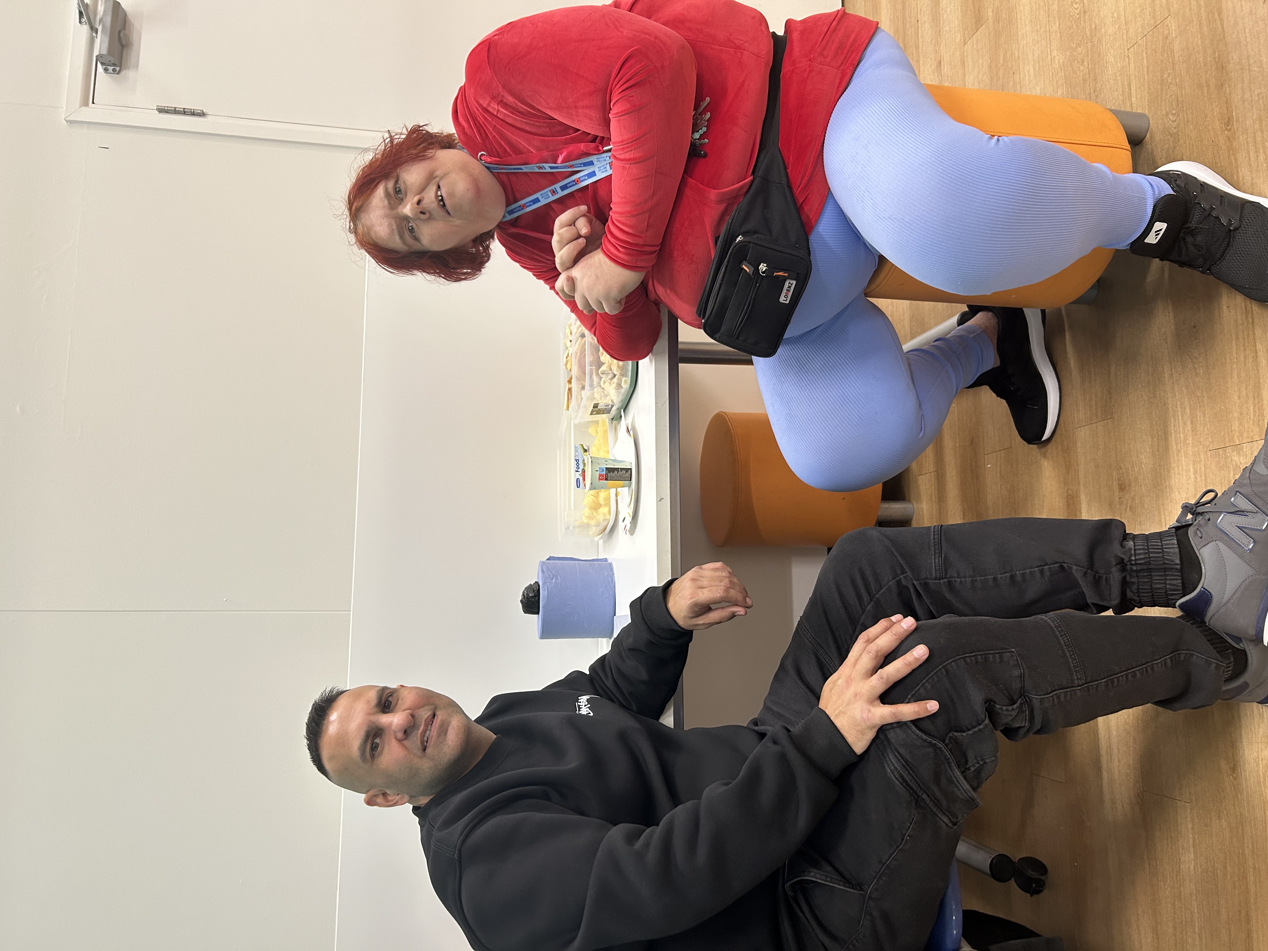 A man in a black jumper and a woman in a red jumper sitting down and smiling at the camera